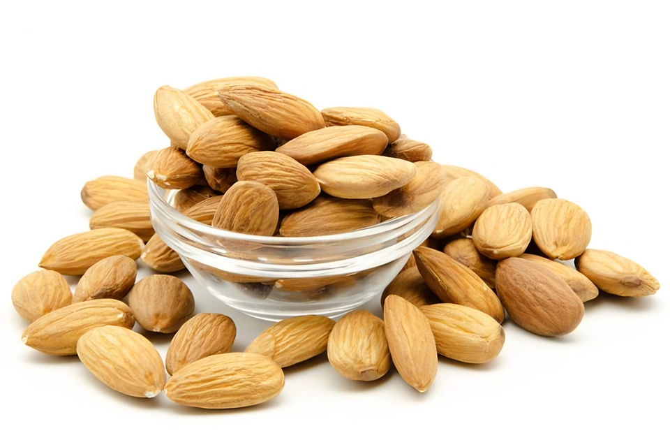 Almendras sin cáscara amontonadas dentro y alrededor de un cuenco de vidrio sobre fondo blanco